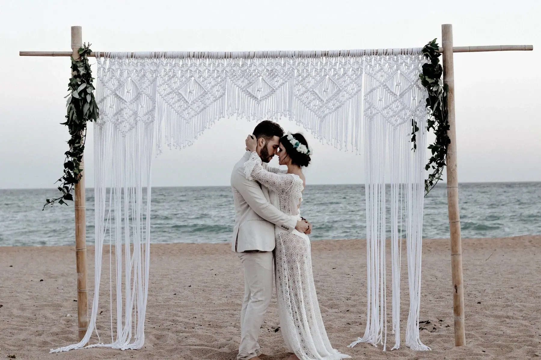 Bride in lace gown and groom in light suit embrace under white macrame beach arch at sunset.