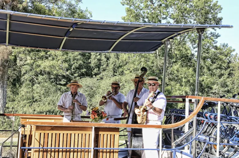 Four white-clad musicians with hats playing trumpet, sax, and trombone on a riverside boat stage under canopy, Burgundy France.