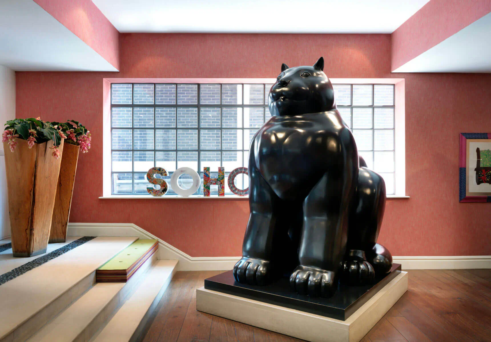 Large black sitting cat sculpture in Soho Hotel lobby with 'SOHO' sign, plants, and pink walls near window.