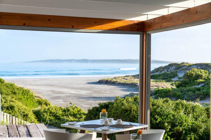 Bright dining area with ocean view of Paternoster beach, dunes, and waves; table set with coffee and vase.