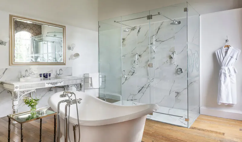 Elegant bathroom at Bailbrook House Hotel with freestanding tub, marble glass shower, white robe, and arched window.