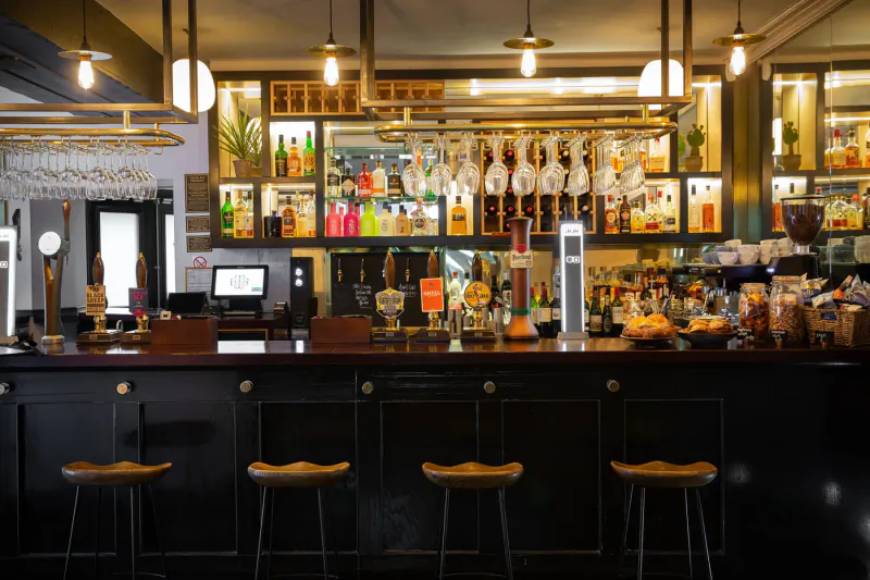 Well-stocked bar at The Black Bull with colorful liquor bottles, hanging glasses, wooden stools, and warm pendant lights