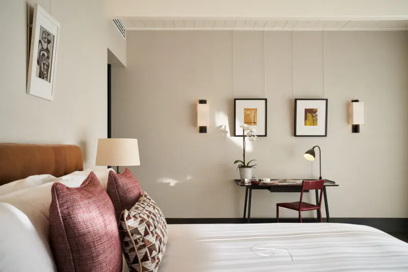 Luxurious hotel bedroom with tan headboard, pink patterned pillows, desk with orchids, and modern art on beige walls.
