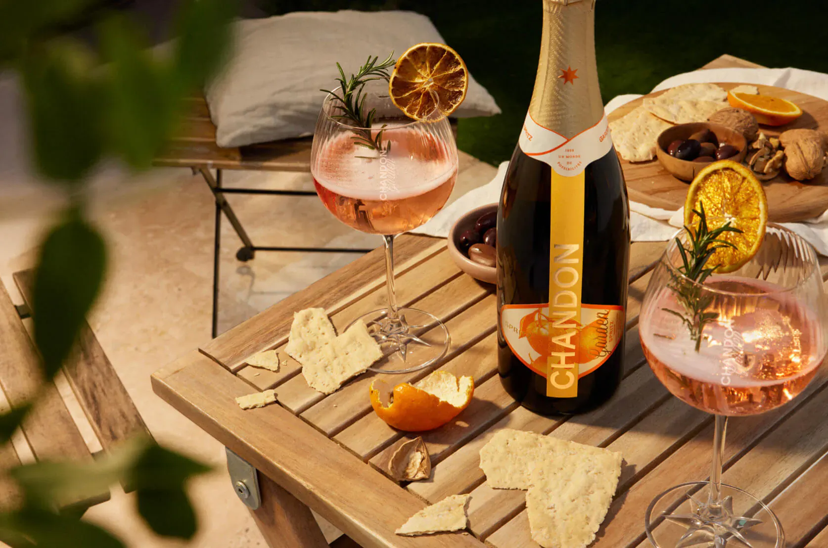 Chandon Garden Spritz rosé sparkling wine bottle with two pink spritz glasses garnished with orange and rosemary on a wooden table outdoors amid snacks.