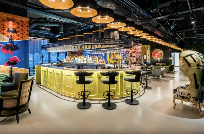 Vibrant punk-inspired bar in nhow London hotel with yellow bar, black stools, colorful murals, and orange pendant lights.