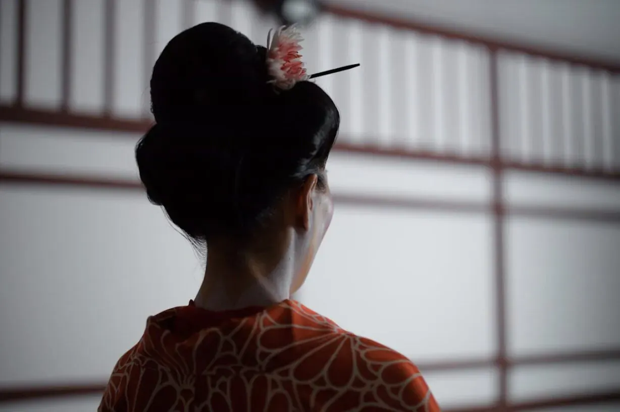 Back view of woman in orange patterned kimono with ornate black updo and pink flower, before shoji screen.