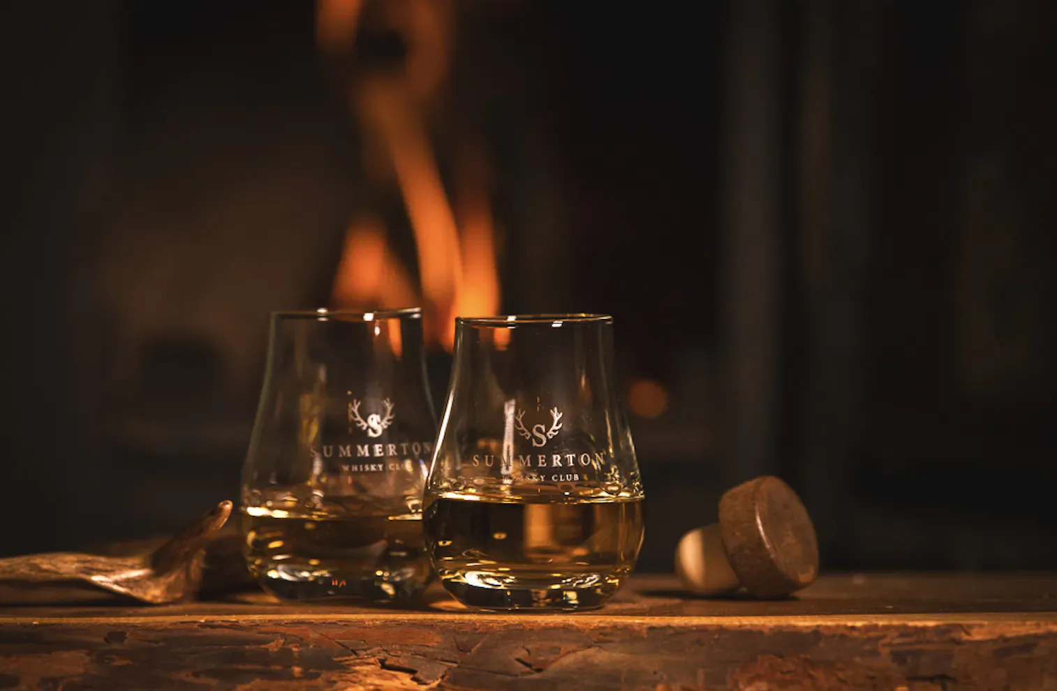 Two Glenturret whisky glasses with amber liquid on wooden table by cozy fireplace, wooden stopper beside.