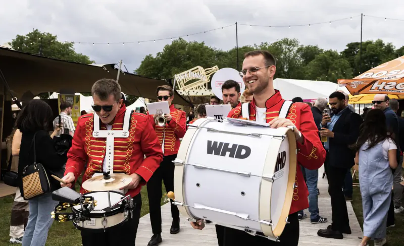 Red-uniformed WHD drummers marching with bass drum and snare at Taste of London event amid tents and crowd.