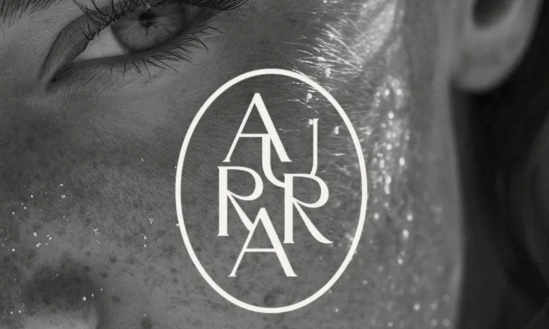 Close-up of woman's eye and cheek with dewy skin, overlaid with 'AURRA' logo, black and white