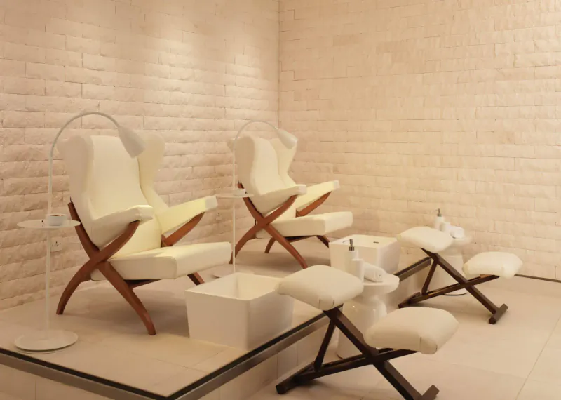 Two white wingback pedicure chairs with footrests and basins in a modern spa with beige brick walls.