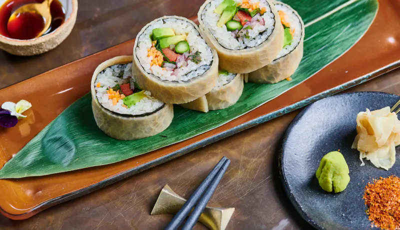 Four colorful veggie sushi rolls on banana leaf platter with chopsticks, wasabi, ginger, and dipping sauce