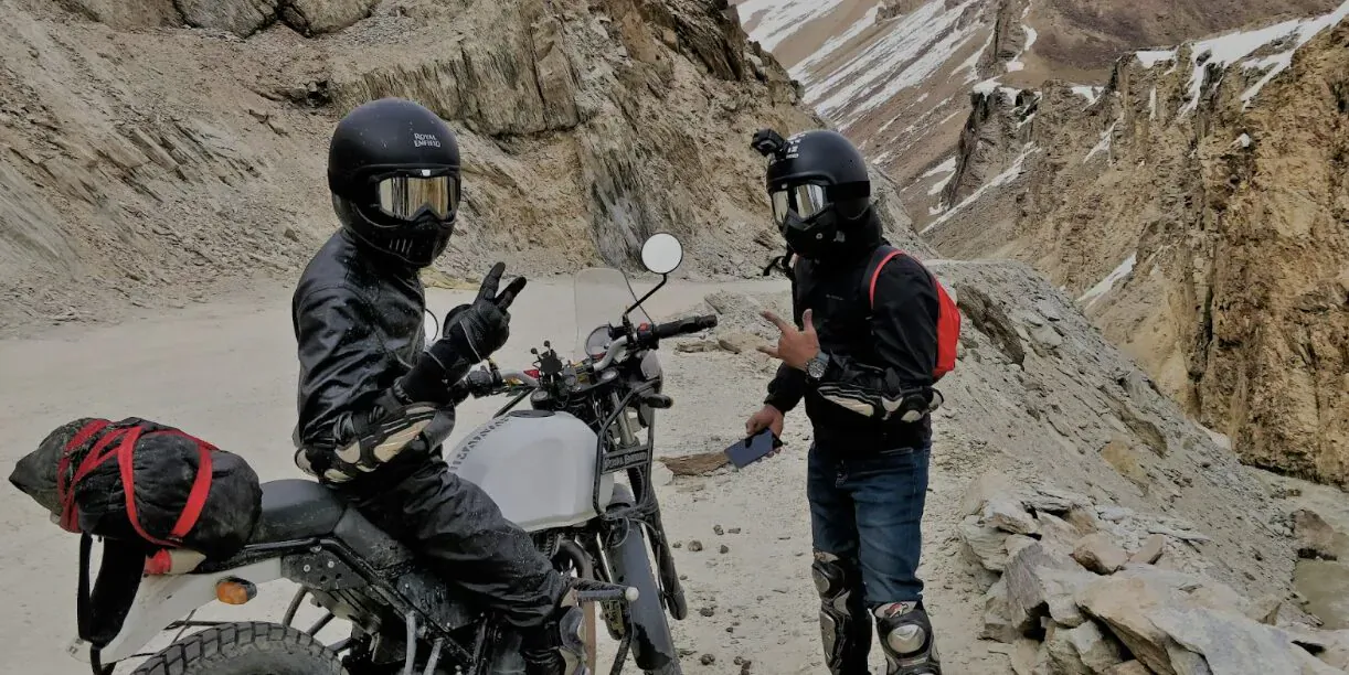 Two helmeted riders in black gear posing with peace signs beside a white motorcycle on a rugged Himalayan mountain road.