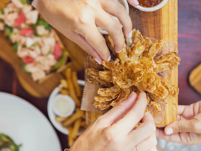 Hands pulling apart a blooming onion on a wooden board with fries and sauces at beachside café.