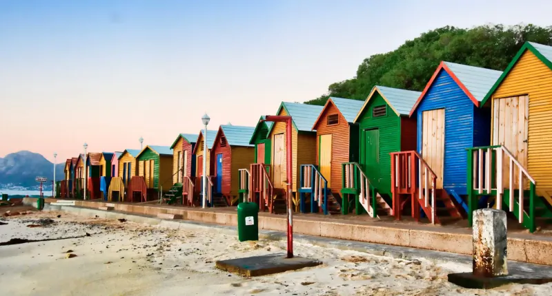 Row of colorful beach huts in vibrant reds, blues, greens along sandy Kalk Bay shore at sunset with mountains.