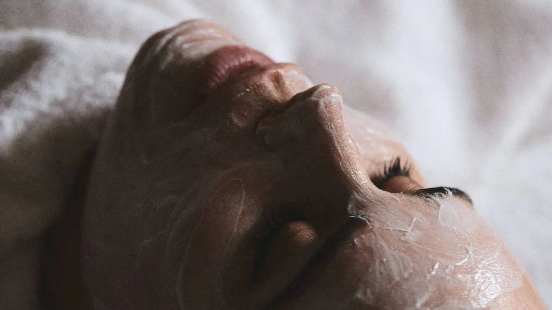 Close-up of woman lying down with white microbiome facial mask on, eyes closed, on white towel