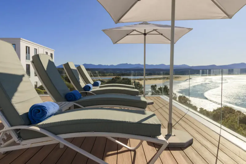 Luxury lounge chairs with blue towels on a sunny oceanfront deck at The Plettenberg Boutique Hotel, mountains in background.