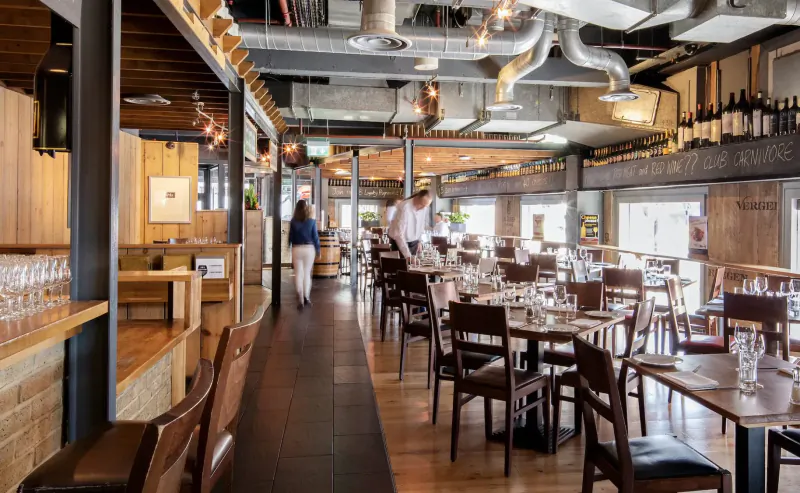 Modern industrial restaurant interior with wooden tables, wine racks, staff walking, and exposed ducts.