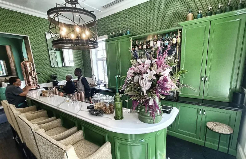 Elegant green bar with emerald cabinets, chandelier, large pink flower arrangement, bottles, and seated patrons.