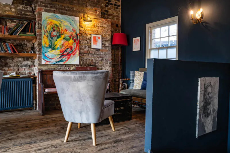 Cozy gallery interior with exposed brick walls, colorful abstract paintings, gray armchair, bookshelves, and blue divider