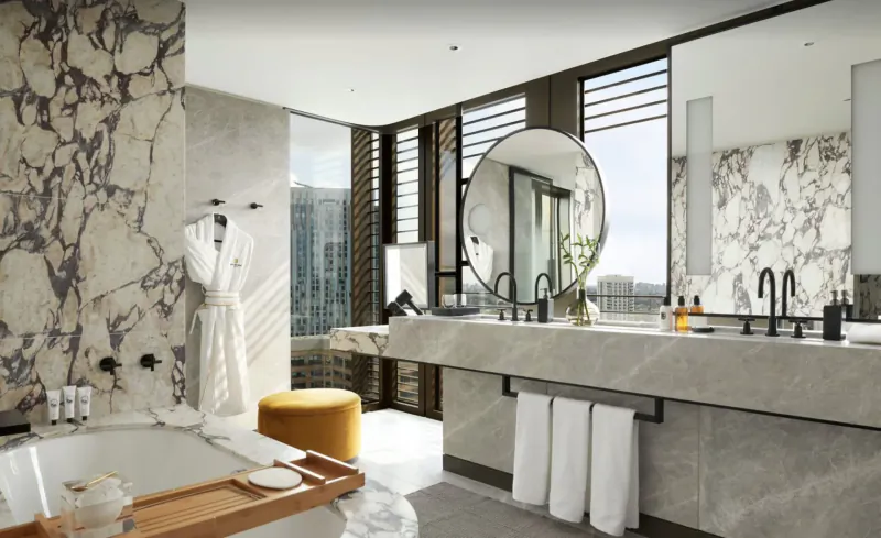 Luxurious modern hotel bathroom with marble walls, freestanding tub, large round mirror, city view, and white robes.