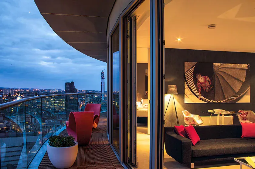 Luxurious Aparthotel Adagio Birmingham balcony at dusk with city skyline view, red chairs, black sofa, abstract art inside.