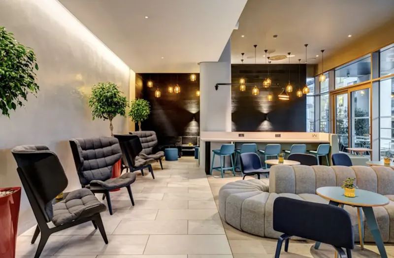 Modern hotel lounge at Apex City of Bath with plush armchairs, curved sofas, bar counter, plants, and warm lighting.