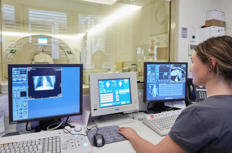 Female radiologist at workstation reviewing MRI and X-ray scans on multiple monitors in medical imaging room
