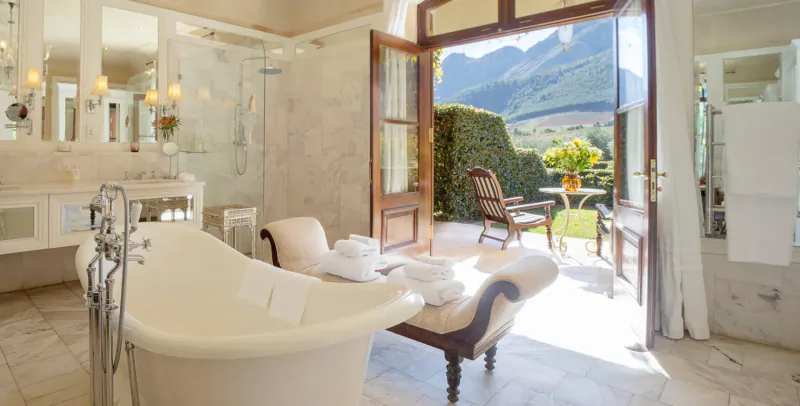 Luxurious white bathroom with clawfoot tub, towels, and open doors to garden and mountain view in Franschhoek.