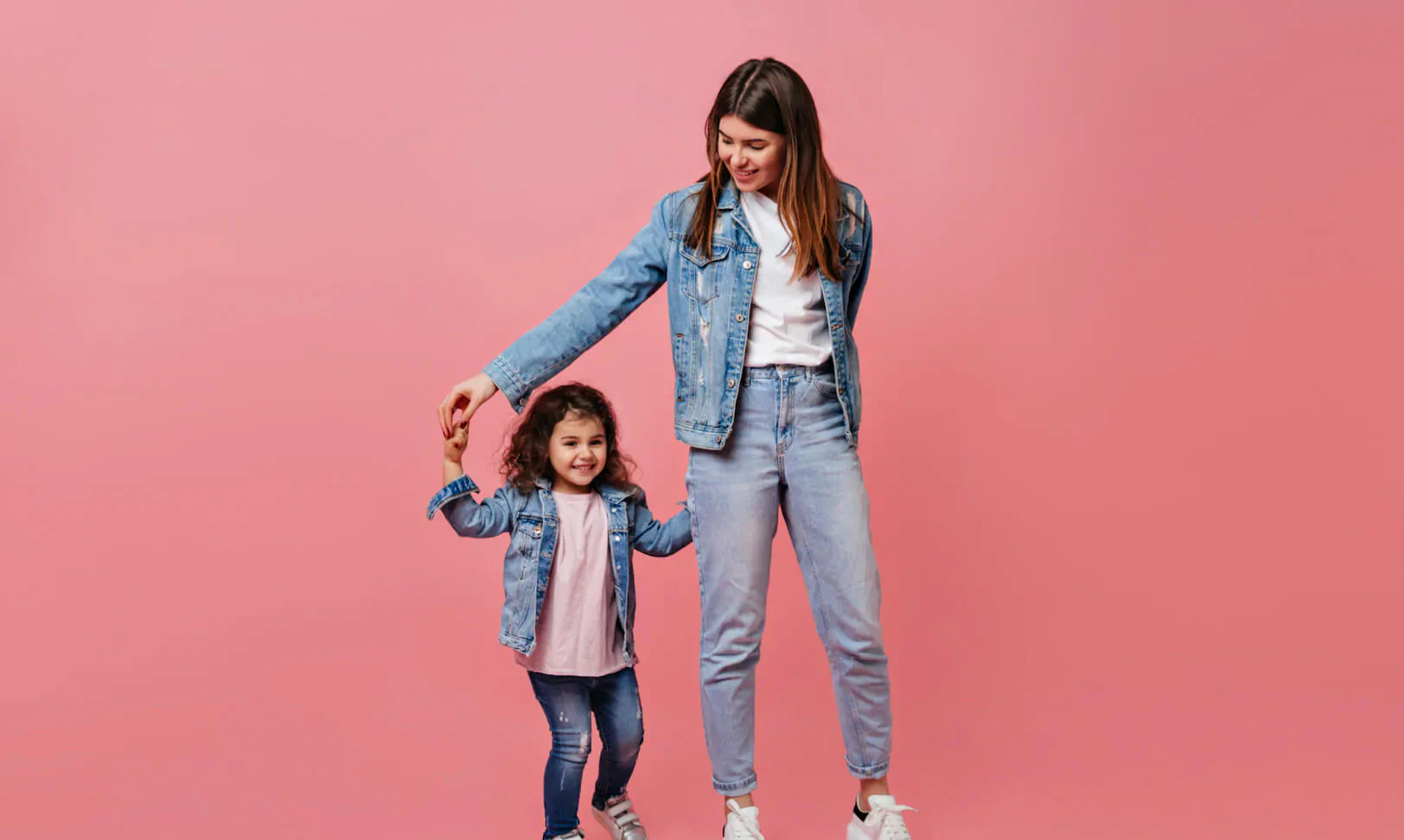 Smiling mother in denim jacket and jeans holds hands with stylish daughter in mini denim jacket, pink background