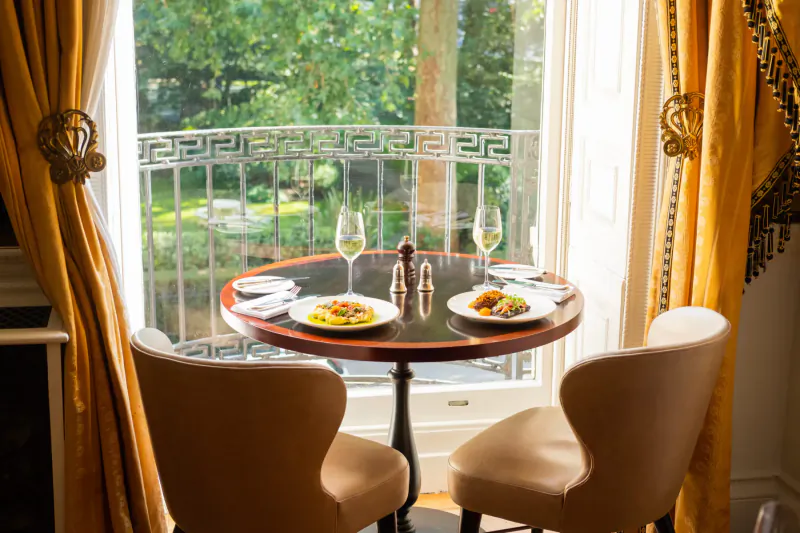 Elegant round table set for two with plates of food, wine glasses, and salt/pepper shakers, by tall window overlooking lush garden in luxurious club.
