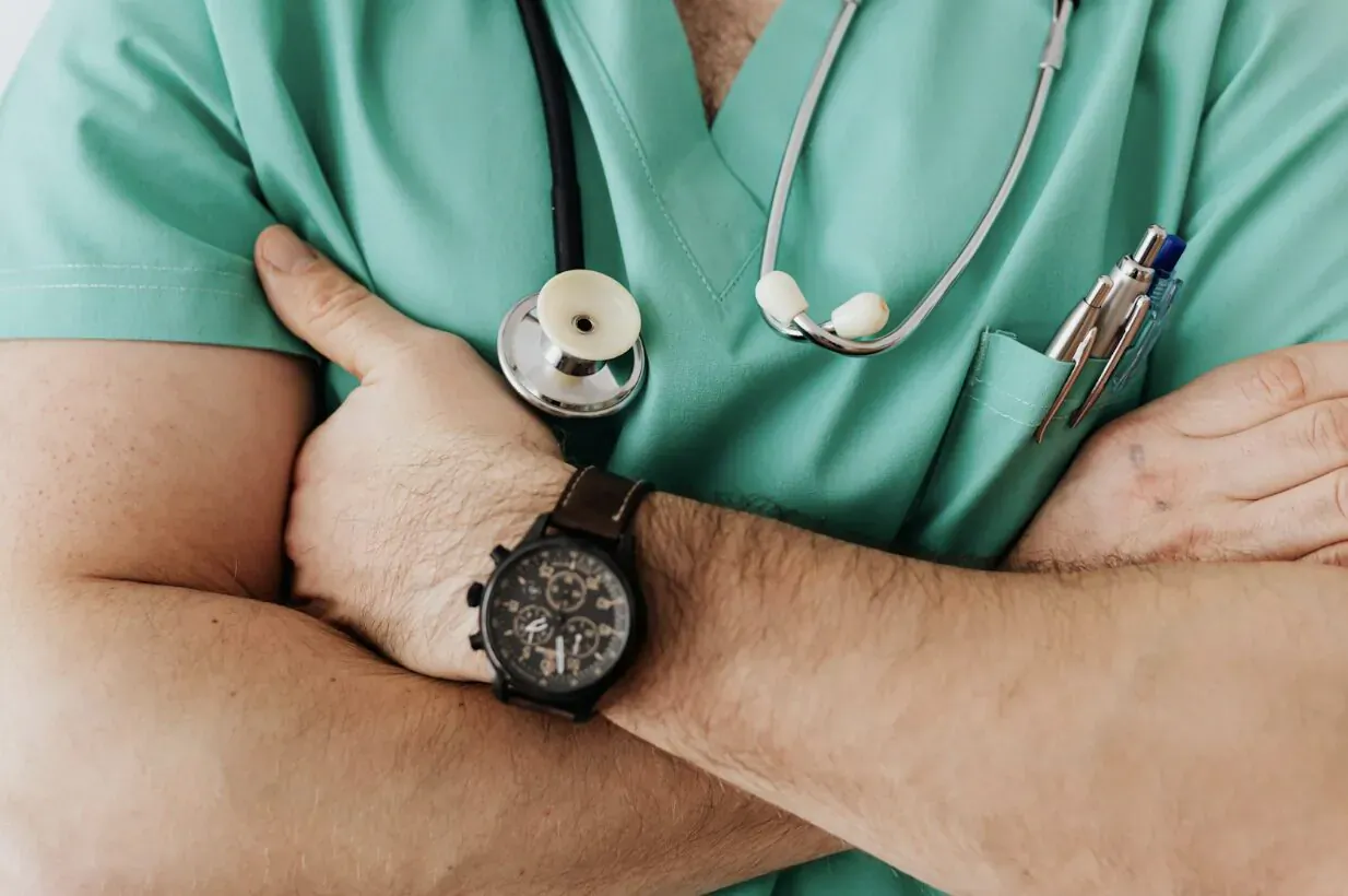 Male doctor in green scrubs with stethoscope around neck, pen in pocket, arms crossed wearing watch