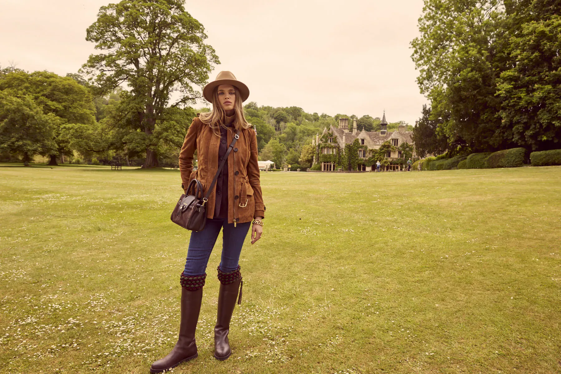 Woman in suede jacket, jeans, riding boots and hat stands on green lawn with manor house and trees behind, Fairfax & Favor style.