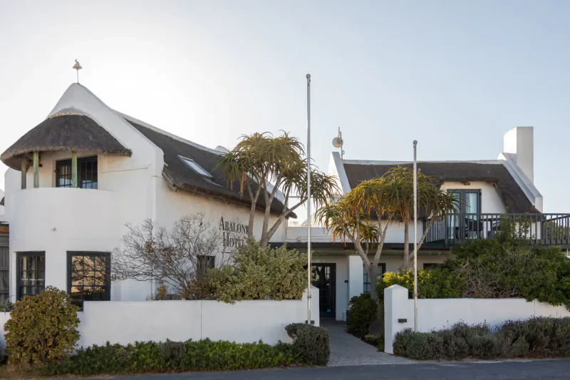 White thatched-roof Abalone Hotel in Paternoster, South Africa, with palms, gardens, and flagpole under blue sky.
