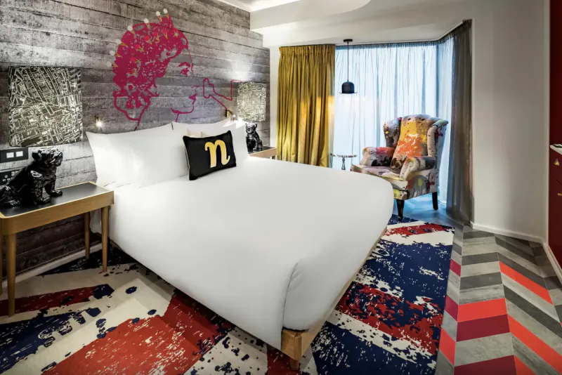 Stylish nhow London hotel room with white bed featuring black 'M' pillow, eclectic armchair, vibrant patterned rug, and punk wall art.