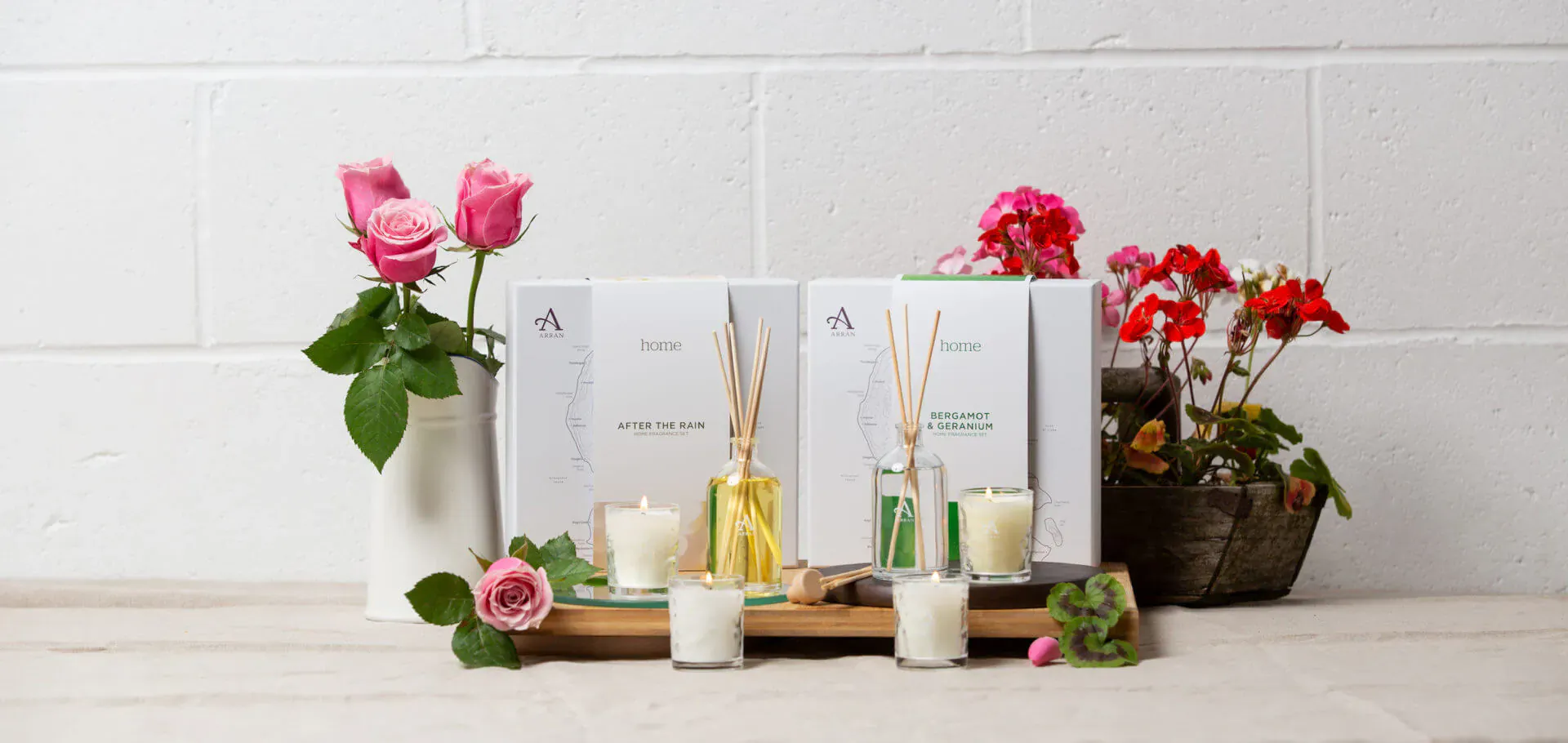 Luxuria Lifestyle reed diffusers, candles, and vases with pink and red geraniums on wooden tray against white brick wall