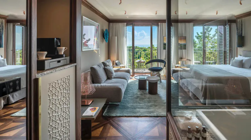 Luxurious hotel suite with gray sofa, bed, bathtub, and panoramic green hill view through large windows