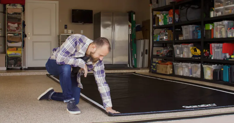 Man kneeling in garage, installing large black GaraMat floor vehicle containment mat.