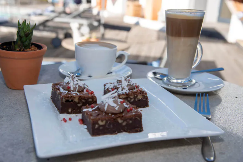 Plate of chocolate brownies topped with coconut and raspberries beside latte and cappuccino on outdoor deck table