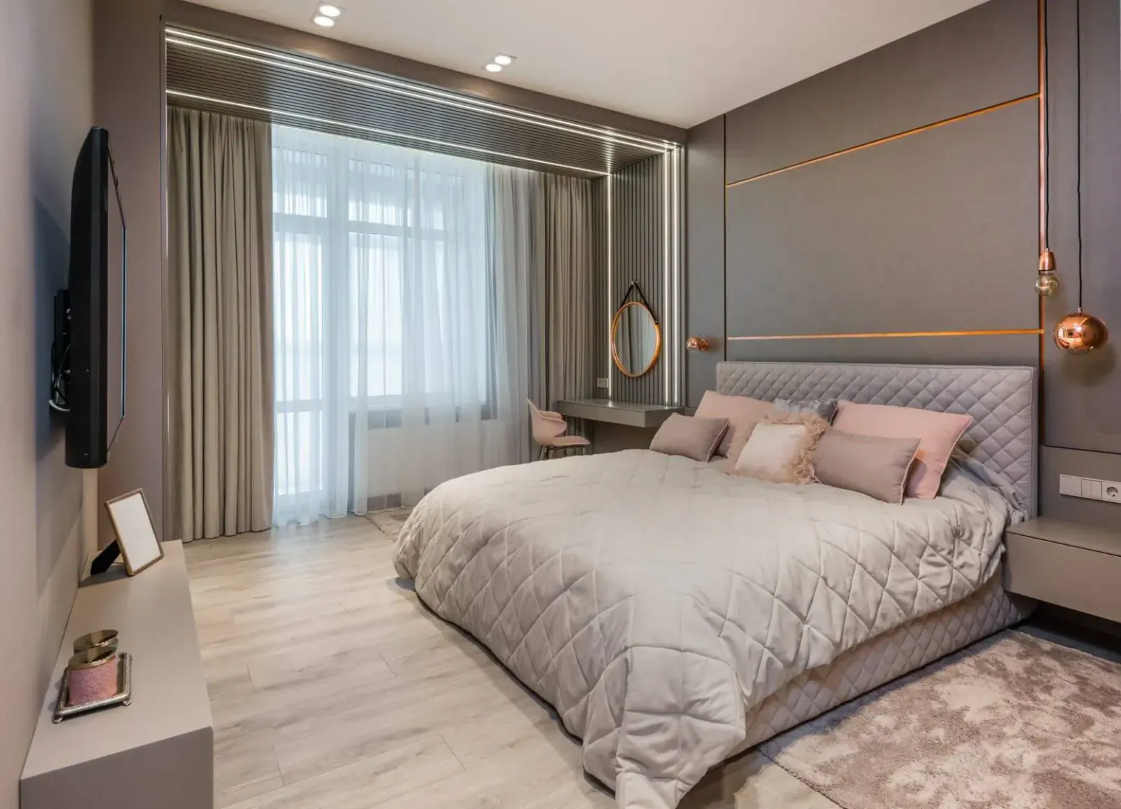 Modern bedroom with gray upholstered king bed, pink pillows, vanity, TV, and large window drapes.