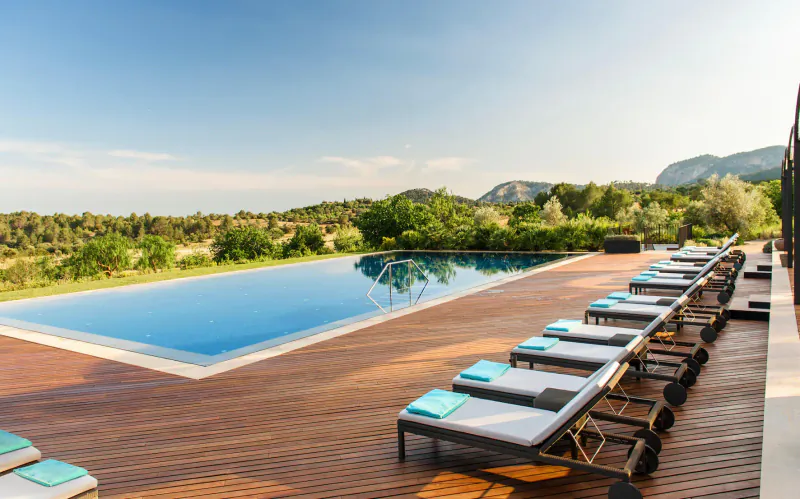 Infinity pool with loungers and towels at Castell Son Claret, Mallorca, overlooking green hills and mountains under blue sky.