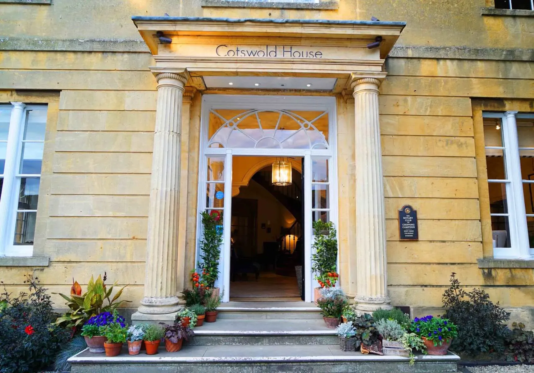 Cotswold House entrance with stone columns, potted plants, and open door revealing interior.