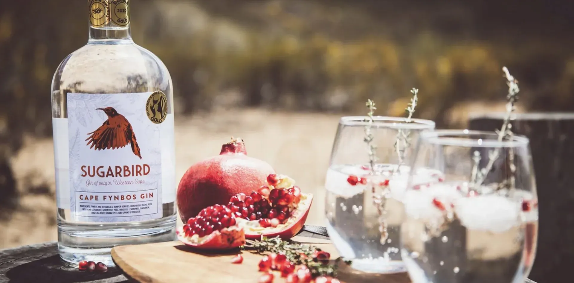 Sugarbird Cape Fynbos Gin bottle with pomegranate slices and garnished gin cocktails on wooden board in outdoor setting