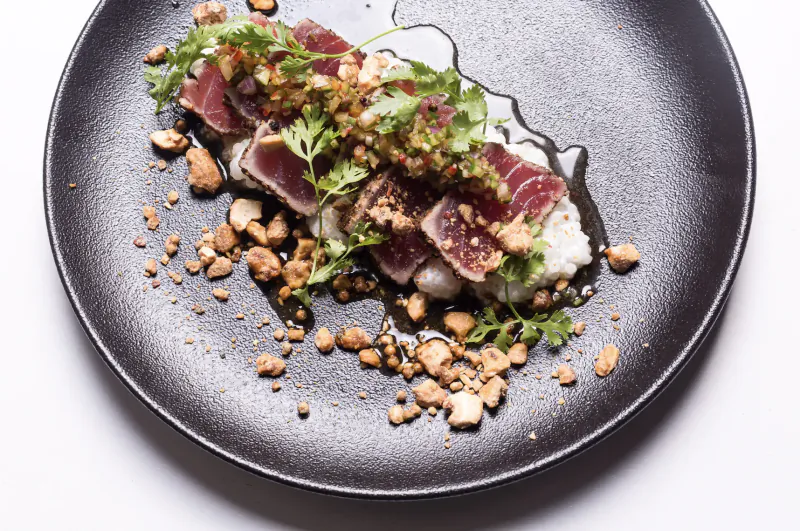 Seared tuna slices with microgreens, white sauce, and crushed nuts on black plate