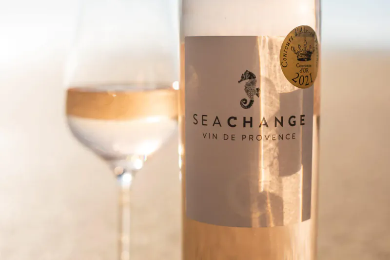 Sea Change Vin de Provence rosé wine bottle with gold award label and poured glass on beach, seahorse logo.