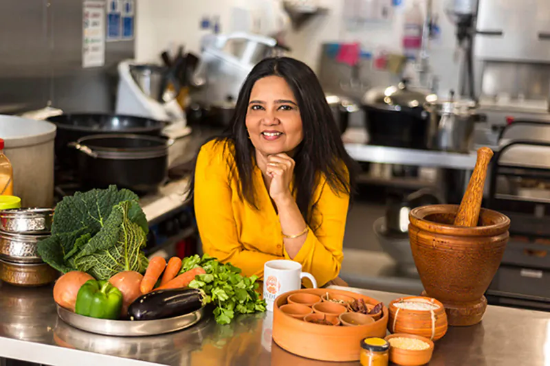 Smiling woman in yellow top poses thoughtfully in kitchen with Indian ingredients, veggies, pots, and mortar pestle on counter