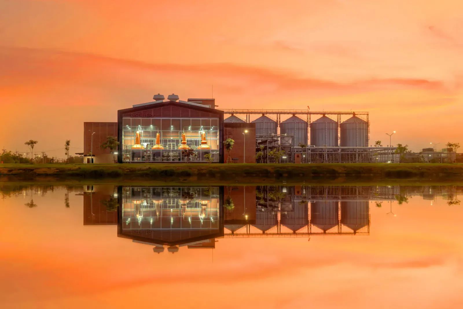 Prakaan Whisky distillery with silos reflected in water at sunset, orange sky.