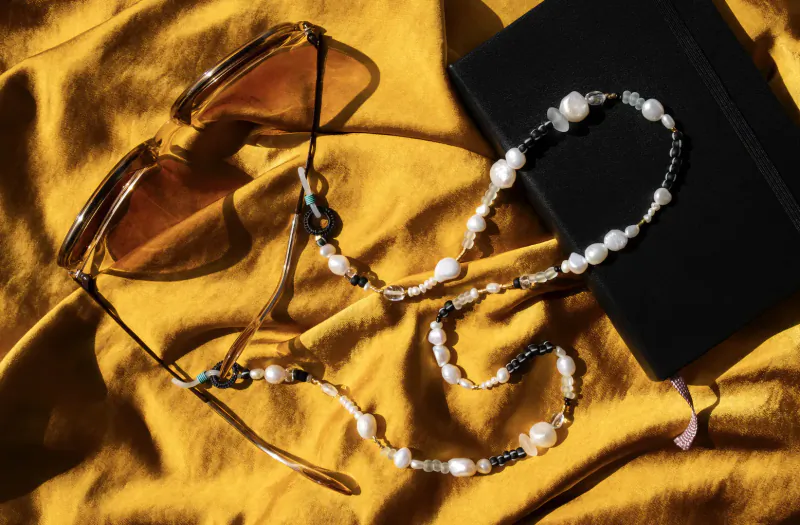 Tortoise-shell sunglasses with white pearl and black bead chain on yellow fabric beside black notebook