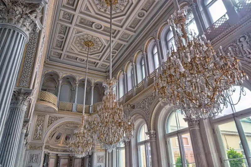 Opulent grand hall with ornate ceiling, marble columns, arched windows, and multiple crystal chandeliers.