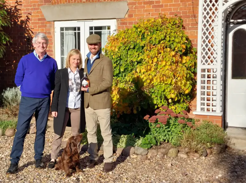 Man in blue sweater, woman in coat, man in tweed jacket with trophy, and brown dog standing outside brick house with yellow bushes.