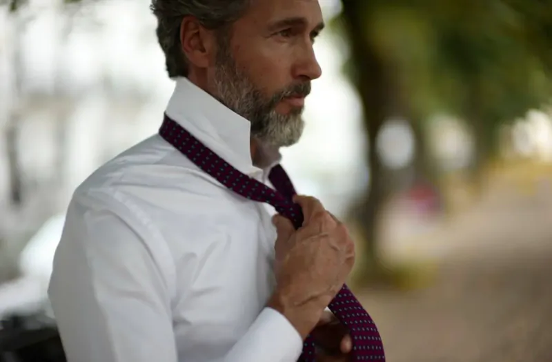 Man with gray beard adjusting purple striped tie on white shirt outdoors near trees
